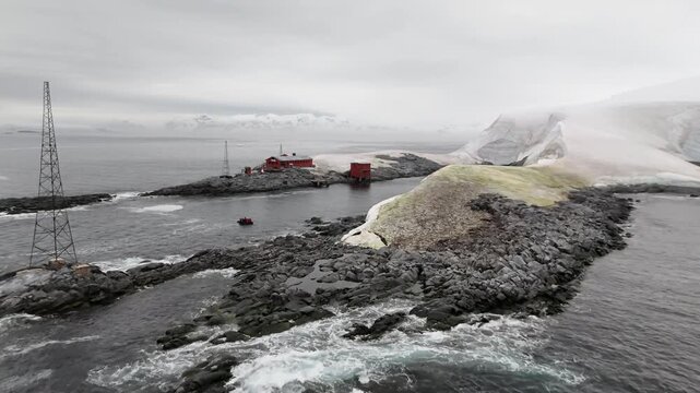 Cinematic aerial flying past a black Zodiac inflatable boat carrying tourists to reveal the red wooden buildings of Argentina's Melchior Base on the rugged volcanic shores of Gamma Island