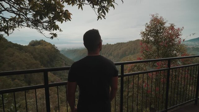 A man stands on a viewpoint overlooking misty forest mountains during early morning. Shot at Tebing Keraton, Bandung, West Java, Indonesia. Concept of solitude, reflection, and connection with nature 