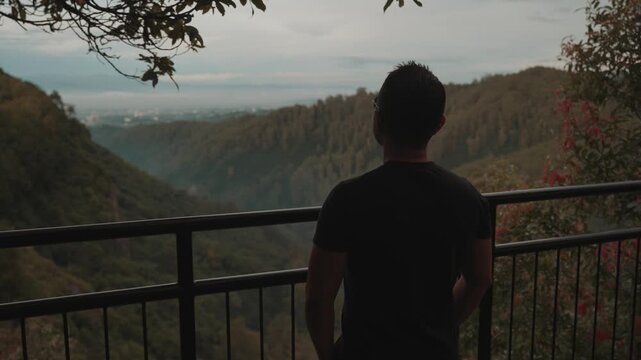 A man stands on a viewpoint overlooking misty forest mountains during early morning. Shot at Tebing Keraton, Bandung, West Java, Indonesia. Concept of solitude, reflection, and connection with nature 