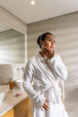 Asian woman wearing bathrobe, standing in bathroom beside vanity and mirror, checking skincare