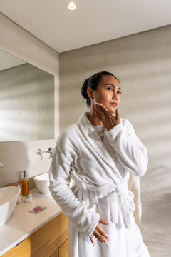 Asian woman wearing bathrobe, standing in bathroom beside vanity and mirror, checking skincare