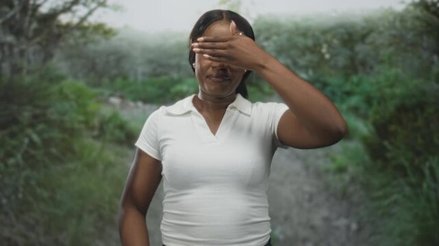 Woman with hand covering face and visible smile, wearing white polo while standing on a forest trail with lush greenery; shy reflection.