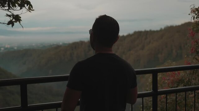 A man stands on a viewpoint overlooking misty forest mountains during early morning. Shot at Tebing Keraton, Bandung, West Java, Indonesia. Concept of solitude, reflection, and connection with nature 