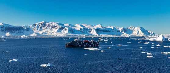 A large dark expedition cruise ship sailing through the icy waters of Antarctica surrounded by icebergs and snowy mountains © NORMA