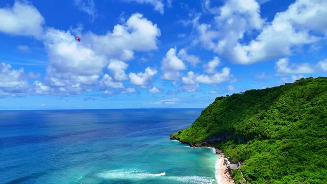 Breathtaking drone footage soaring over turquoise ocean waters along dramatic limestone cliffs in Uluwatu, Bali. Lush green hills and rugged coastline with crashing waves. Captured in 16:9 horizontal 