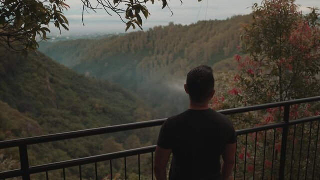 A man stands on a viewpoint overlooking misty forest mountains during early morning. Shot at Tebing Keraton, Bandung, West Java, Indonesia. Concept of solitude, reflection, and connection with nature 