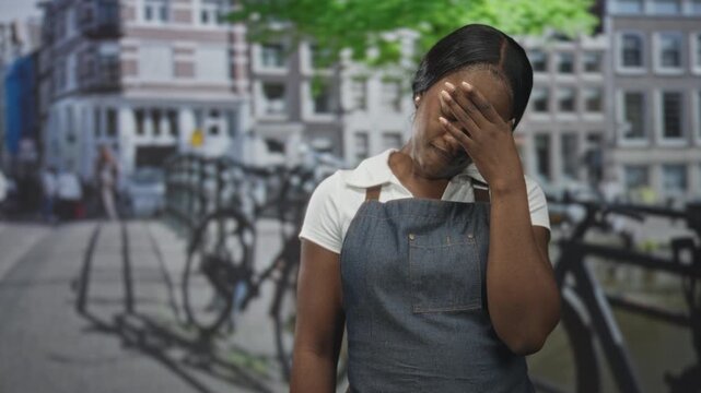 Woman in apron covers face with hand on street by canal and bicycle rack near buildings, visible covering gesture; fatigue duty.