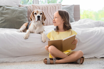 Obraz premium Cute little girl with Beagle dog reading book near bed in bedroom