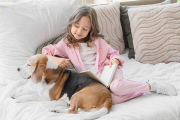 Obraz premium Cute little girl with Beagle dog reading book on bed in bedroom