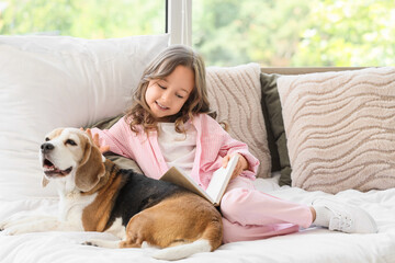Obraz premium Cute little girl with Beagle dog reading book on bed in bedroom