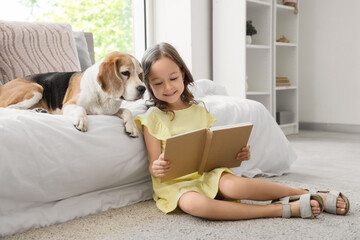 Obraz premium Cute little girl with Beagle dog reading book near bed in bedroom
