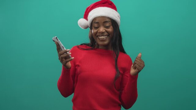Woman holding phone with thumbsup gesture wearing santa hat and red sweater in studio; holiday celebration joy.