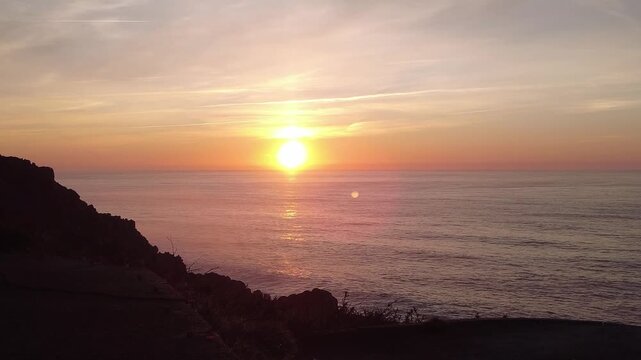 Golden sun slowly setting over the calm ocean horizon, casting a warm glow on the water and silhouetting the rocky coastline of prior cape in a breathtaking, serene natural display