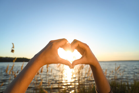 Female hands making heart gesture against river at sunset. Summer solstice