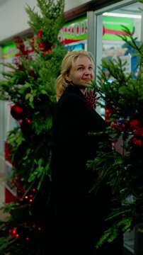 Joyful woman in a black winter coat pauses to touch a snowflake ornament on a decorated tree before entering a local shop at night. Authentic holiday lifestyle footage.