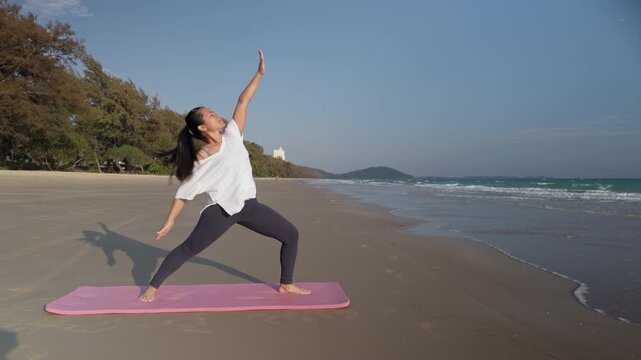 Asian woman practicing yoga reverse warrior pose on tropical beach in Thailand. Outdoor fitness and wellness scene ideal for travel, health and lifestyle content.
