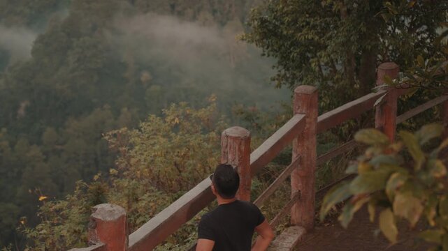 A man stands on a viewpoint overlooking misty forest mountains during early morning. Shot at Tebing Keraton, Bandung, West Java, Indonesia. Concept of solitude, reflection, and connection with nature 