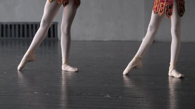 Close-up of two ballet dancers in pointe shoes. Legs move gracefully on dark wooden floor. Red patterned skirts visible. Background shows plain studio walls