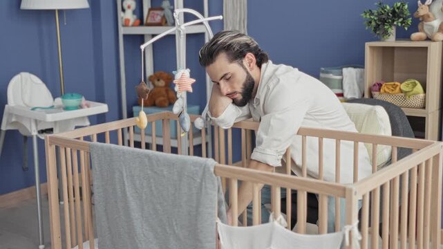 Man leans over wooden crib, rests head on hand and reaches into cradle in a nursery building; fatigue parenting.