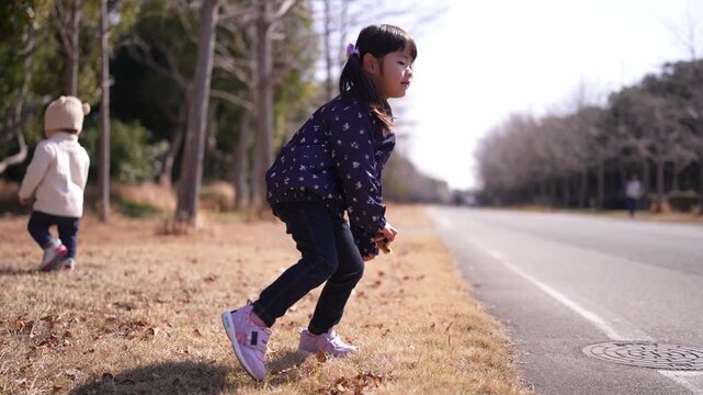 Two children play near a quiet road. One sits, the other walks past. Sunlight glints off bare trees. Quiet, sunny moment captured. Perfect for family or outdoor scenes. Children wear casual