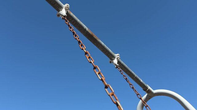 Weathered, rusty swing chains creak as they sway back and forth. The orange-brown oxidized metal creates a stark, textured contrast against the deep, cloudless blue of a wide Midwestern sky.