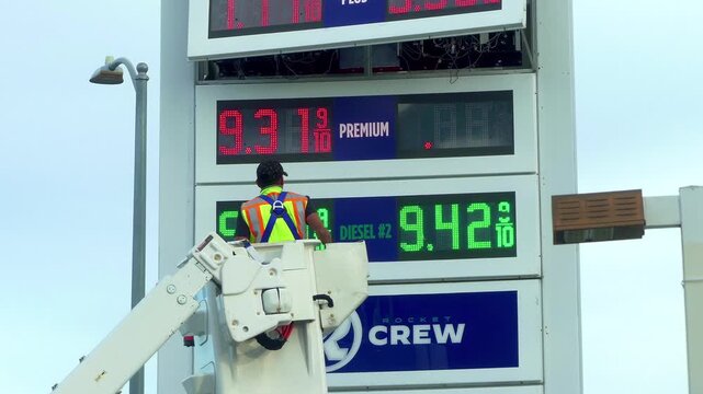 A worker in a bucket lift updates and services an illuminated gasoline price digital sign showing prices above $6 per gallon for regular fuel at a gas station in Los Angeles, California