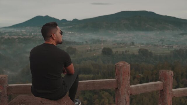 A man stands on a viewpoint overlooking misty forest mountains during early morning. Shot at Tebing Keraton, Bandung, West Java, Indonesia. Concept of solitude, reflection, and connection with nature 