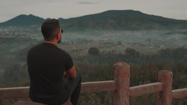 A man stands on a viewpoint overlooking misty forest mountains during early morning. Shot at Tebing Keraton, Bandung, West Java, Indonesia. Concept of solitude, reflection, and connection with nature 