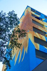Colorful mural facade on modern apartment building, abstract sunflower pattern, blue yellow shapes rising against sky expressing urban art creativity architecture identity, positive city atmosphere © ullision