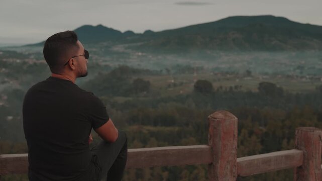 A man stands on a viewpoint overlooking misty forest mountains during early morning. Shot at Tebing Keraton, Bandung, West Java, Indonesia. Concept of solitude, reflection, and connection with nature 