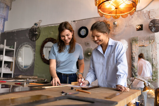 Craftswomen colleagues collaborating on glasswork decor, sketching design, planning project, working together on glass sconce stand at workbench table with material. Teamwork, craftsmanship, artisanry