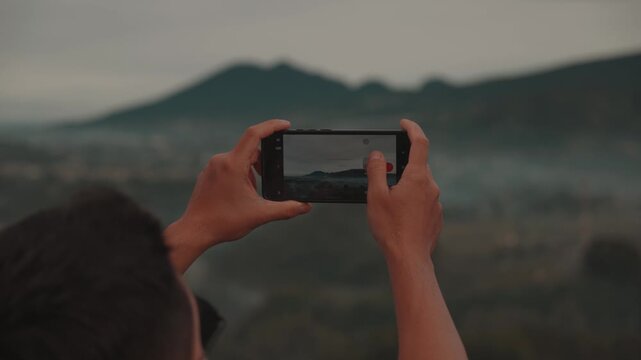 Traveler using smartphone to take photos and selfies with a misty mountain forest landscape. Filmed at Tebing Keraton, Bandung, West Java, Indonesia. Concept of modern travel, social media, and captur