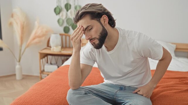 Young hispanic man sitting crosslegged on bed with hand to forehead and other hand on hip showing headache in building; fatigue reflection.