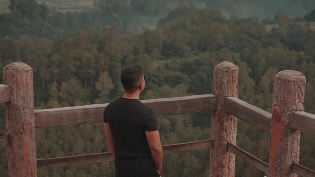 A man stands on a viewpoint overlooking misty forest mountains during early morning. Shot at Tebing Keraton, Bandung, West Java, Indonesia. Concept of solitude, reflection, and connection with nature 