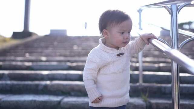 Cute toddler grips metal railing, staring downward. Sunlight glows behind stone steps. Child wears soft, cream-colored sweater. Curious expression, eyes focused on ground. Perfect for parenting