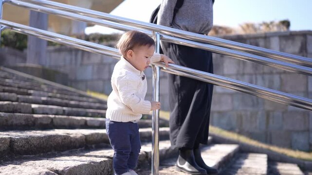 Little toddler grips rail while climbing stone steps. Child looks down, curious, focused on next step. Adult stands nearby, guiding but not helping. Outdoor scene suggests family outing, calm