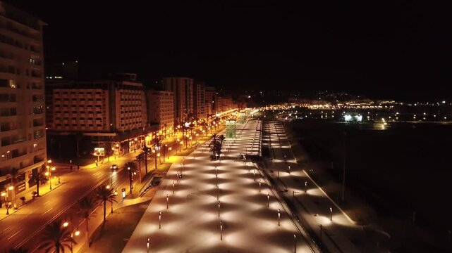 Aerial drone view of Tangier waterfront promenade at night with illuminated walkway, palm lined boulevard and city lights along the Moroccan coast.