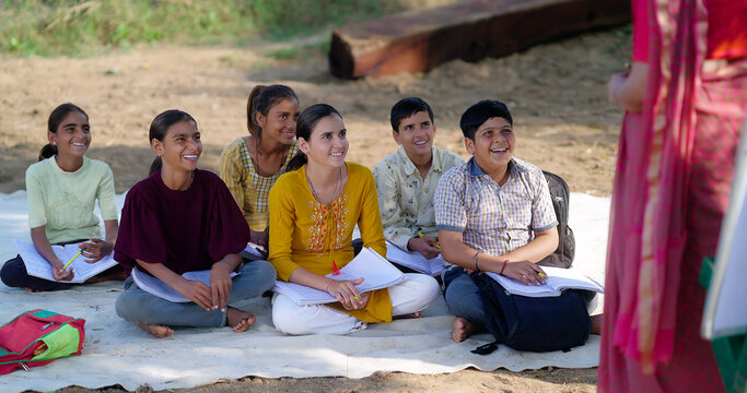 Indian mature adult gen z female lady talk speak share good news teach to villager group day under tree. Happy girl boy sitting on mat do study look board learn at open class field rural area place