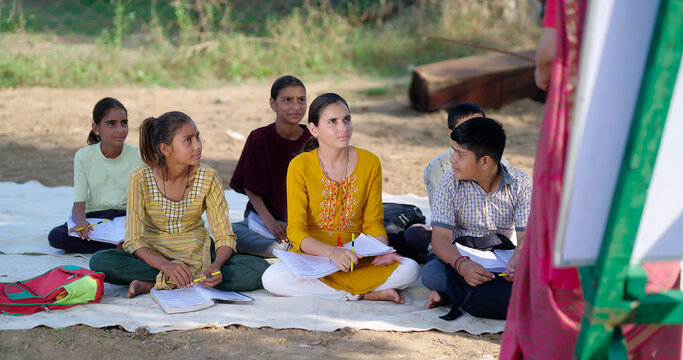 Indian mature adult gen z female lady talk speak share good news teach to villager group day under tree. Happy girl boy sitting on mat do study look board learn at open class field rural area place