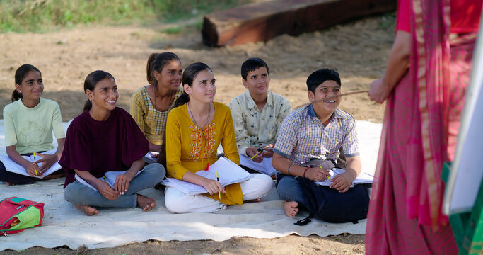 Indian mature adult gen z female lady talk speak share good news teach to villager group day under tree. Happy girl boy sitting on mat do study look board learn at open class field rural area place