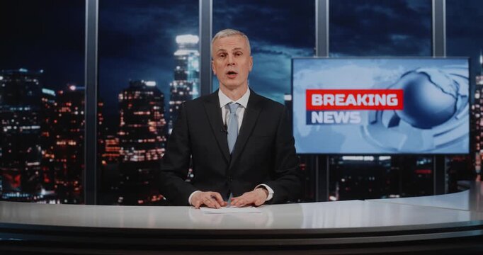 Professional Mature Male Anchor in Dark Suit and Blue Tie Sits at Desk, Looking at Camera and Talking. Concept of Television Journalism in Studio With Breaking News Screen and Night City View.