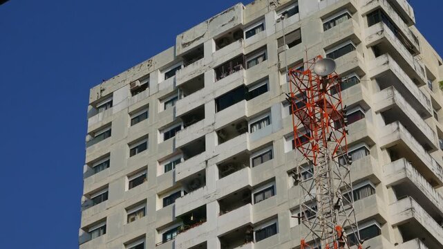 Tall white concrete residential high-rise building beside an orange lattice telecom transmission tower against a vivid blue sky