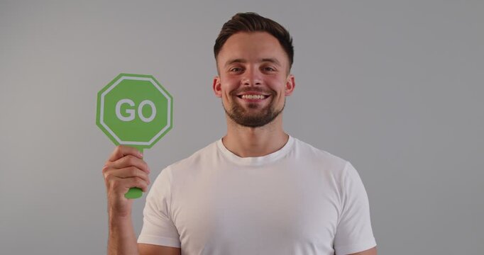 Smiling man holds two green GO traffic signs. The man is holding octagon paddles with the word go, showing a smile and gesture of approval, set against a gray studio background with copyspace.