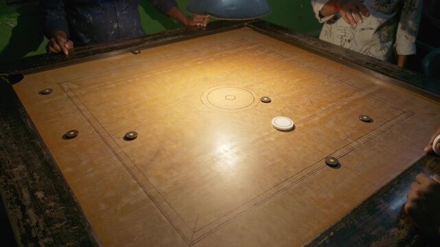 Players around carrom board, striker sliding across wooden surface with coins spread out, active gameplay under warm light in indoor night setting