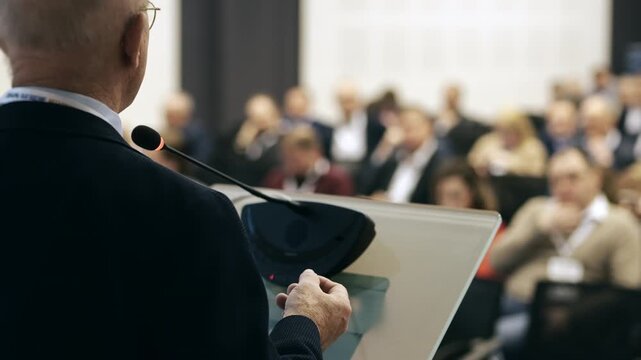 Business speaker talking to audience during conference presentation from podium with engaged listeners in background indoor event setting.