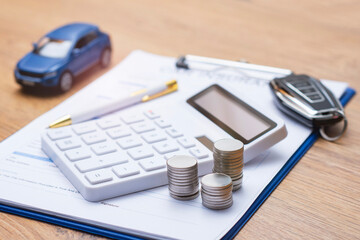 Obraz premium Car insurance document on clipboard with white calculator, stacked coins, blue toy car and modern car key on wooden desk. Concept auto insurance premium calculation and vehicle finance planning.