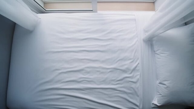 Overhead view of a bed in a bedroom with an open window, a light breeze billowing the curtain, concept of promised coolness for an afternoon rest