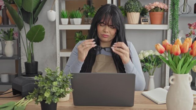 Woman florist at laptop wearing apron and stretching arms behind head in flower studio with tulips on table; work fatigue.