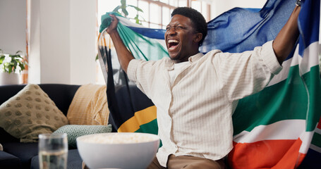 Black man, South African flag and celebration in home, soccer and cheering for champion. Fan, shouting and happy person in lounge, football and excited for score lead, sports tournament or supporter © peopleimages.com