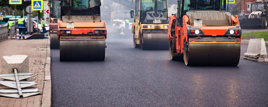 Large machines are paving asphalt on a city street. Workers are present observing the process. Traffic signs and barriers are visible as the work is ongoing during daylight hours.
